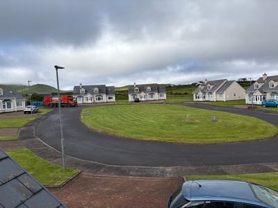This is a medium-angle, outdoor, daytime shot of a residential street in Portmagee, Ireland. The sky is overcast and appears to be a bit stormy. The street is paved and curves around a circular patch of bright green grass. Several houses, each with grey roofs and light-colored walls, are visible on either side of the street. They appear to be duplexes or townhouses. A red garbage truck with blue bins is parked on the left side of the street. A silver car is parked in front of one of the houses on the left, and a blue car is parked further down the street on the right. A lamppost stands tall in the foreground, near the curve of the road. The landscape behind the houses is hilly and green, suggesting a rural or coastal setting. The overall impression is of a quiet, overcast day in a residential neighborhood.