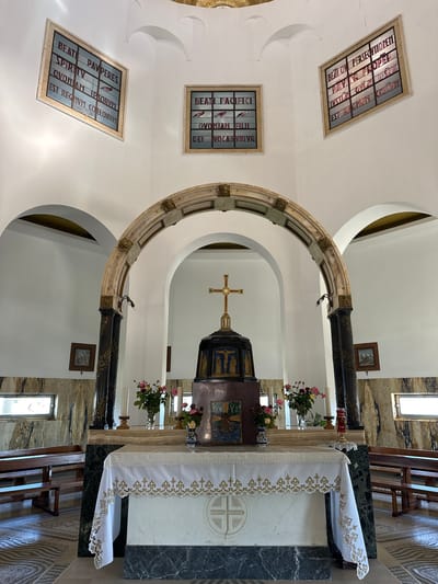 The image depicts the interior of a chapel, likely the Mount of Beatitudes in Emek HaYarden Regional Council, Israel. In the foreground, an altar is draped with a white tablecloth adorned with gold embroidery and a cross motif. Behind the altar, a structure with a crucifix on top is visible, framed by a marble arch supported by dark columns. Stained glass windows with religious text and imagery are mounted on the walls. Benches for worshipers are arranged on either side of the chapel, and the floor is tiled in a mosaic pattern. The walls are adorned with marble panels and small framed pictures. Natural light streams in from windows, illuminating the space.