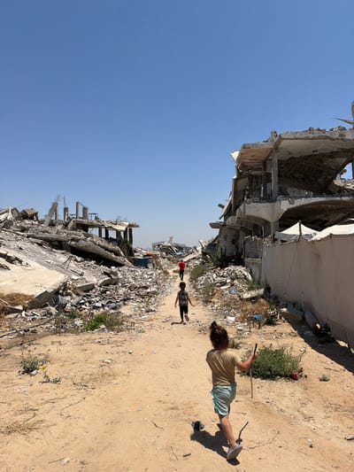 The image depicts a street scene in Jabalia, Palestinian Territories, characterized by widespread destruction. In the foreground, a young child with dark hair pulled back in a ponytail is walking away from the camera, wearing a light brown t-shirt and light blue shorts, holding a stick. Further down the dusty path, another child in a dark t-shirt and dark pants is also walking away, and a third child in a red shirt is visible in the distance. The environment is dominated by the remnants of bombed-out buildings, with concrete debris, rebar, and rubble filling the landscape. The sky is clear and blue. The presence of children navigating this devastated area suggests resilience amidst destruction.