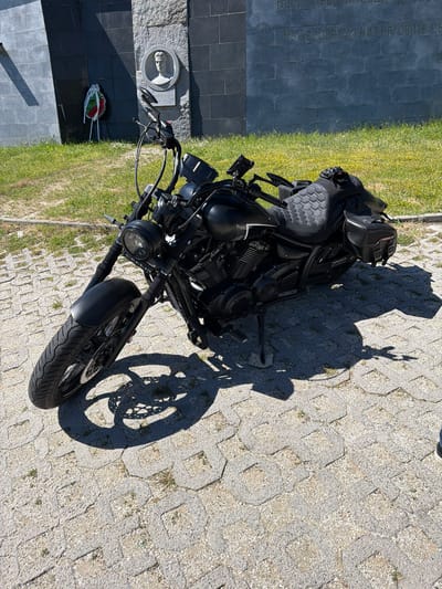 A motorcycle is parked on a paved surface adjacent to a grassy area in Bunovo, Bulgaria. The motorcycle is black and has saddlebags. Next to the motorcycle is a monument or memorial. The shadow of a person and motorcycle is cast on the paved surface.