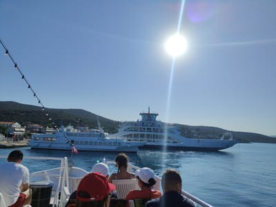 The image depicts a sunny day at sea, likely in the Municipal Unit of Pelasgia, Greece, given the architectural style and mountainous terrain visible. The foreground shows the rear view of several people seated on a white ferry. A man in a white t-shirt and red shorts is on the far left, facing away from the camera. Next to him, a woman with dark hair is seated, looking towards the water. Two children, one wearing a red cap and the other a white cap, are also visible in the foreground.
In the middle ground, two large white ferries are docked or maneuvering in the water. The ferry closer to the foreground has the name "VENUS" visible on its side. The ferry further back appears larger and more modern. A red and white flag flies from a pole on the closer ferry. The water is calm and reflects the bright sunlight.
The background consists of a green, mountainous coastline with several buildings clustered along the shore, indicative of a coastal town or village. The sky is a clear, bright blue, with the sun prominently visible in the upper right quadrant, causing a lens flare. A string of lights is visible along a railing in the upper left portion of the image. The overall scene suggests a leisurely ferry journey on a warm day.