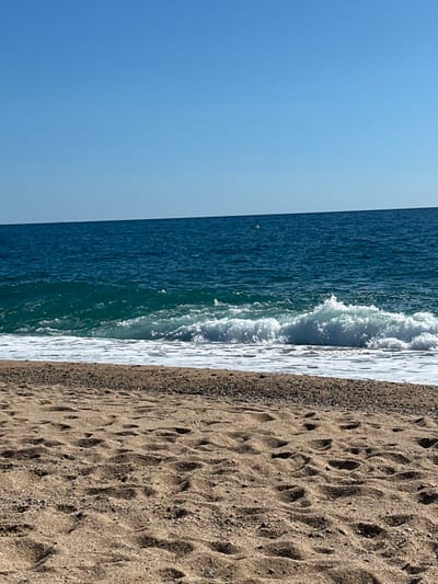 The image depicts a beach scene in Pineda de Mar, Spain, under a clear, bright blue sky. The foreground consists of tan-colored sand marked with numerous footprints and small, dark pebbles along the water's edge. In the midground, the turquoise ocean water exhibits breaking waves with prominent white foam. Further out, the sea transitions to a deep blue, with a single yellow spherical buoy visible on the surface, positioned slightly right of the frame's center. The horizon line, where the sea meets the sky, is distinct. No human figures, animals, or man-made structures other than the buoy are present in the frame.
