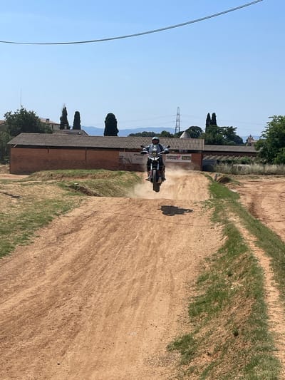 A person is riding a motorcycle on a dirt track in L'Ametlla del Vallès, Spain. The motorcycle is kicking up dust as it moves across the track. The rider is wearing protective gear. The environment consists of a dirt track with grassy edges, a building with a sloped roof in the background, trees, and an overhead electrical power line. There are mountains in the distant background under a clear blue sky. The scene is in daylight with the sun casting shadows.