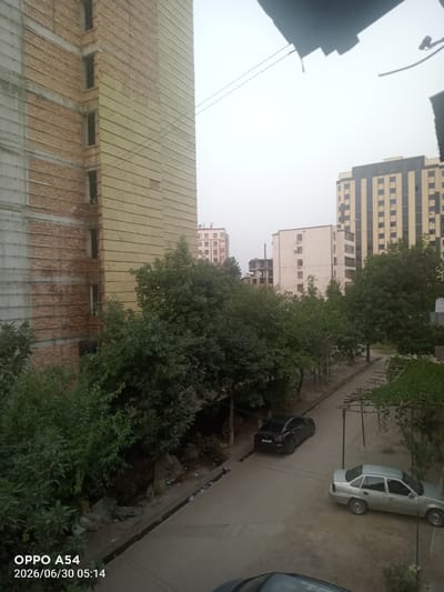 The image shows an urban street scene in Bokhtar, Tajikistan. The foreground features a partially constructed building with exposed brickwork and insulation, suggesting ongoing construction or renovation. Mature trees line a paved street, casting shadows and creating a shaded environment. Several cars are parked along the street, including a black sedan and a silver sedan. In the background, multiple apartment buildings of varying architectural styles are visible. One building is tall and modern with a striped facade, while others are smaller and appear to be older. The sky is overcast, indicating a cloudy or hazy day, and the lighting suggests it is either early morning or late afternoon. The overall atmosphere is quiet and residential, with no people visible in the frame. The timestamp "2026/06/30 05:14" and the text "OPPO A54" are overlaid on the bottom left corner of the image, likely indicating the device and date the photo was taken.