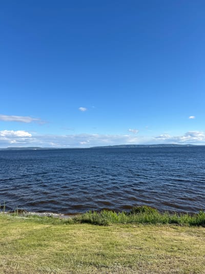 The image depicts a serene natural landscape. The foreground features a grassy slope leading down to the edge of a large body of dark blue water. The water is slightly rippled, suggesting a gentle breeze. In the mid-ground, across the water, a long, low-lying landmass is visible, characterized by rolling hills or mountains. The sky above is a clear, bright blue, punctuated by a few scattered white clouds, particularly on the left side of the frame. There are no people or man-made structures immediately visible in the image, emphasizing the natural setting. The scene appears to be a calm, sunny day. This image could potentially be of the Volga River near the city of Primorsky, Russia, given the vast expanse of water and the distant, hilly shoreline typical of that region.