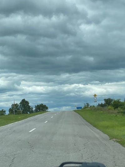 A grey, overcast sky looms over a paved road that ascends a gentle incline. White lines mark the lanes of the road, which appears somewhat worn and cracked. To the right of the road, lush green grass borders the asphalt, and a scattering of trees, predominantly green and deciduous, dot the landscape. In the distance, several road signs are visible. One sign, a yellow diamond with a black border, is accompanied by a white triangle with a black border pointing downwards, and below that, a rectangular sign with white text on a white background. Further to the left, a blue rectangular sign is positioned near a cluster of trees. The scene suggests a rural or semi-rural setting, potentially on the outskirts of a town or village. There are no people or animals visible in the image.