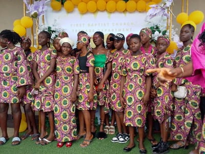 A group of teenage girls stand in front of a wall decorated with gold balloons and a sign that reads "First Baptist Church, Teenage Department". They are all wearing matching pink and green patterned dresses. Some girls are holding small purses. One girl is wearing a black hat with a white inscription. The girls are all smiling and looking at the camera. The scene is likely a gathering of the First Baptist Church's youth group. The coordinates provided locate the scene in Yenagoa, Bayelsa State, Nigeria.