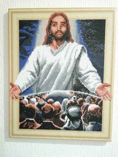 A framed diamond painting depicts Jesus with outstretched arms facing a crowd. The painting is hung on a textured white wall, likely in a residential setting. The overall scene is static and indoor. The painting itself is composed of small, faceted beads, giving it a shimmering appearance. The background of the painting features a dark, starry sky transitioning to a lighter, possibly celestial glow behind Jesus. Below Jesus, a multitude of figures are depicted, rendered in shades of brown, gray, and white, suggesting a dense gathering of people. Jesus is centrally positioned, wearing a white robe, and his facial features are detailed with brown hair and beard. His arms are extended outward, palms up, in a gesture of welcome or blessing towards the assembled crowd. The diamond painting is enclosed in a light-colored frame. The surrounding wall has a subtle, raised pattern. No specific location in посёлок опытного хозяйства «Ермолино», Russia can be determined from the provided image.
