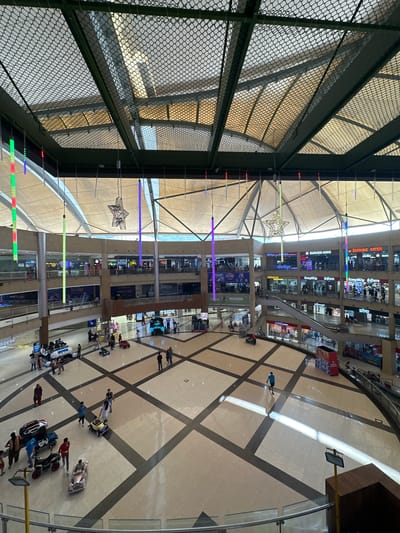Here's a detailed description of the image:
The image shows the interior of a multi-level shopping mall in Kalyan-Dombivli, India, viewed from an elevated position looking down into the central atrium. The atrium floor is tiled with a patterned design. Several people can be seen walking around and operating ride-on toy cars.
The surrounding levels of the mall are lined with shops and restaurants. Signage for various businesses such as "Burger King," "Easy Buy," and "Barbeque Nation" are visible. There are lights suspended from the ceiling, some arranged in vertical lines, and a decorative star-shaped light fixture.
A section of the view is obstructed by a wire mesh, suggesting that the photograph was taken from behind a protective barrier, possibly a balcony or a high walkway.
