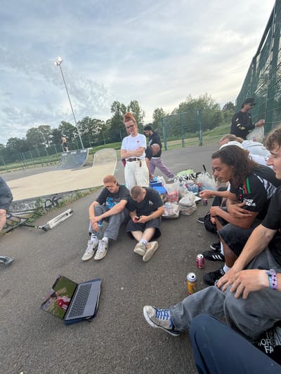 A group of approximately twelve people are gathered near a skatepark at an outdoor location in Kettering, United Kingdom. The scene appears to be taken in the daytime under a cloudy sky. The skatepark features a ramp. A light pole is visible. The ground is asphalt. The group is a mixture of males and females, mostly young adults. Some are sitting or lying on the ground. There are several bags, cans, bottles, and a cooler. There is a laptop displaying a scene, possibly a video. A scooter is on the ground near the skatepark. The group appears to be relaxing, some in physical contact, possibly resting or in conversation.