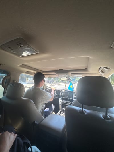 A man is driving a car on a sunny day in Jezqazğan, Kazakhstan. He is wearing a light-colored polo shirt and dark pants, and his hands are on the steering wheel. The interior of the car is visible, including the dashboard, seats, and a sun visor. A small flag, likely the flag of Kazakhstan, is hanging from the rearview mirror. The exterior of the car shows a street with buildings and trees. The overall scene depicts a typical day of transportation within the city.