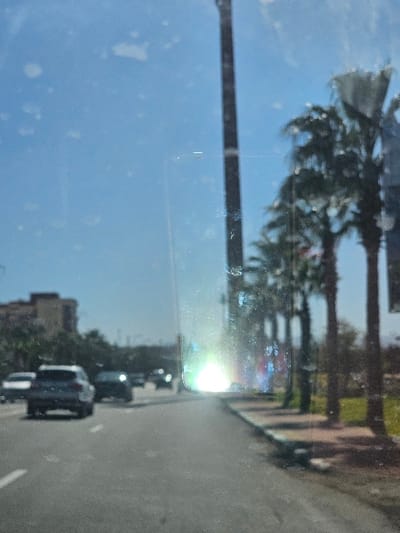 The image presents a daylight street scene in Boufakrane, Morocco, captured from an internal vehicle perspective through a smudged or dirty windshield. A clear blue sky is visible above. The foreground consists of a paved asphalt road with white lane markings. Several vehicles are present on the road, traveling away from the camera; a light-colored SUV is most clearly discernible in the left foreground.

To the right of the roadway, a sidewalk composed of reddish-brown pavers runs parallel to the street. A row of tall palm trees lines the sidewalk, with green ground cover visible beyond them. A tall, dark, slender vertical structure, consistent with a monopole or antenna, rises prominently on the right side of the image, extending upwards out of frame.

On the left side of the road, multi-story buildings are visible in the distance, partially obscured by green vegetation. A strong light flare, characterized by an intense white glow and surrounding prismatic colors, is present in the center-right of the image, obscuring details in that area. No individuals or specific interactions are identifiable.
