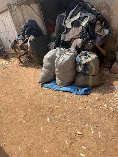 The scene depicts an outdoor commercial or storage area in Sabon Gari South, Nigeria. The foreground consists of reddish-brown dirt ground with scattered small debris including wood fragments and white paper pieces. In the midground, five filled sacks are positioned on a crumpled blue tarp. Three larger, light-grey woven sacks stand upright, with two smaller, similarly woven sacks, one featuring a green stripe, stacked behind them. Behind these sacks, a visible alcove entrance of a light-colored, weathered building reveals two large piles of assorted garments on wooden tables or stands. A larger garment pile is situated within the building's alcove, and faint graffiti is visible on the wall above this pile. To the far left, a white wooden door with vertical panels is partially visible, partially obscuring a section of a dark-colored vehicle, consistent with a motorcycle or scooter. No individuals are present in the frame.