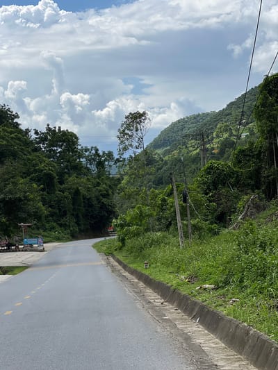 This image depicts a scenic view of a road winding through a lush, green, mountainous landscape, likely in the area of Thuận Hòa Commune, Vietnam. The road is paved and has dashed yellow lines marking the lanes. To the left of the road, there is a concrete area with a small stall or cart displaying signs and some items, possibly a roadside vendor. Further left, a few trees and a blue and white sign are visible.
The right side of the road features a concrete curb and a steep embankment covered in dense, green vegetation and tall grasses. Several utility poles with electrical wires run along the roadside, interspersed with the abundant greenery. In the background, a forested mountainside rises steeply, with scattered buildings visible amongst the trees. The sky above is partly cloudy, with large, white cumulus clouds against a blue sky, suggesting a daytime scene, possibly after or before a rainfall. The overall impression is one of natural beauty and rural tranquility.