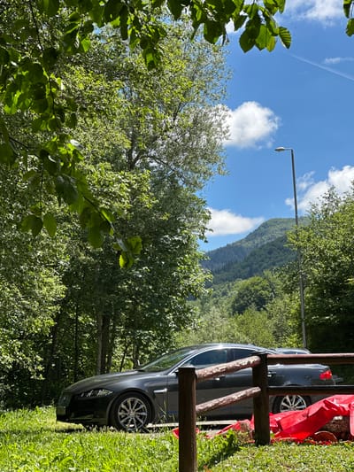 An eye-level shot captures a dark gray Jaguar sedan parked on a grassy area beside a rustic wooden fence. The car is angled towards the right, with its front left wheel and part of the driver's side visible. Behind the car, a dense forest of green trees stretches out, with a prominent, tall streetlamp positioned to the right of the vehicle. In the background, a majestic, tree-covered mountain rises under a clear blue sky dotted with fluffy white clouds. A thin white contrail is visible in the upper right portion of the sky. The scene is bathed in bright sunlight, casting shadows and highlighting the vibrant green of the foliage. The location is identified as Bare Kraljske, Montenegro.