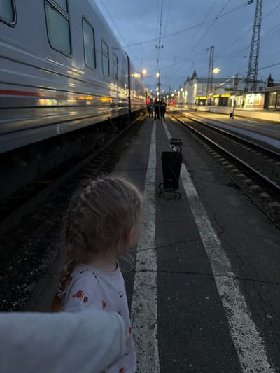 The image captures a scene at the Rtishchevo railway station in Russia at dusk. A young girl with blonde hair in braids, wearing a pajama top with a print, stands on the platform. A train is adjacent to the platform. Other people are further down the platform. Overhead wires and streetlights are visible. The ground is paved with white lines. A black metal trashcan sits on the platform.