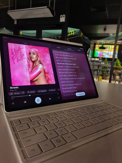 In Chuchuligovo, Bulgaria, a tablet displays a music application with the song "Streets" by Doja Cat playing. The screen shows album art featuring the artist in pink, lyrics, and playback controls. The tablet is placed on a white keyboard accessory, suggesting a workspace or entertainment setup. In the blurred background, a McDonald's restaurant's exterior with awnings and an illuminated sign is visible, indicating the location is likely near or within a commercial area. The time displayed on the tablet is 02:47.