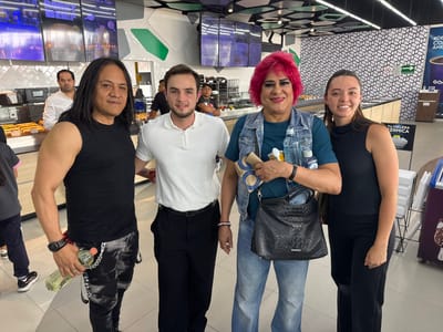 Four individuals pose for a group photo inside a modern convenience store or bakery, identified by "Gomart" branding and "Panadería" signage, located in San Buenaventura, Mexico. All four subjects are smiling cheerfully at the camera, suggesting a friendly and casual interaction.

On the far left, a man with long, dark hair and a black sleeveless top holds a clear plastic bottle with a yellowish liquid. He wears black and white patterned pants adorned with chains. Beside him, a man with short dark hair and a beard is dressed in a white polo shirt and black trousers. To their right, a person with vibrant pink/red hair, heavy makeup, and a denim vest over a teal top wears light blue jeans. They hold a black textured handbag, a paper bag, and a water bottle. On the far right, a young woman with dark hair wears a black sleeveless top and black wide-leg pants.

The background reveals a commercial setting with a counter displaying baked goods such as hot dogs in buns and various breads. Overhead digital screens show food items and pricing. The store's decor includes white tiled walls, a distinctive black-and-white geometric pattern on another wall, and green accents. Visible Spanish text includes "Panadería" (Bakery), "MASA MADRE" (Sourdough), "ROAST BEEF," "De la Casa" (Of the House), and "Por solo $35.00." Other text seen on displays and signs includes "Los Pioles," "TU HIELERA TÉRMICA" (Your Thermal Cooler), and a "RUTA DE EVACUACIÓN" (Evacuation Route) sign, confirming the Mexican context. The bright lighting suggests an indoor daytime setting.