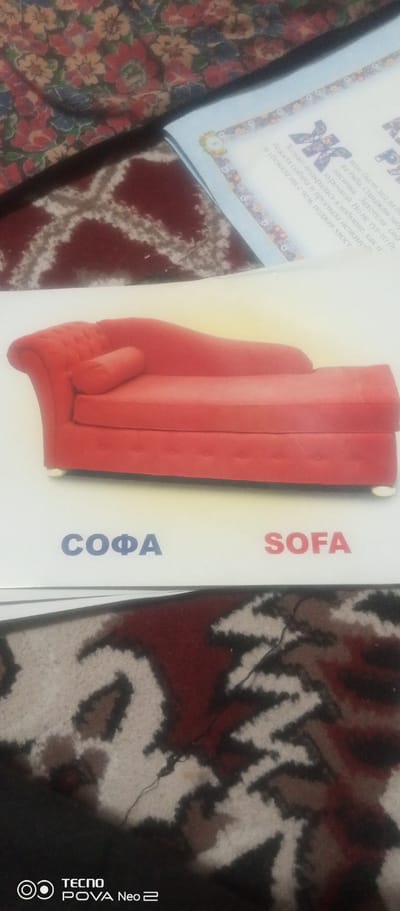 The image shows a close-up, vertical shot of an object that appears to be a red chaise lounge, positioned on a white surface. Below the chaise lounge, the words "СОФА" are written in blue Cyrillic font on the left, and "SOFA" is written in red English font on the right. The background consists of a patterned fabric, likely a rug or upholstery, with dark red and beige/white colors. The lighting is somewhat uneven, casting shadows. The lower portion of the image shows a black area with a TECNO POVA Neo 2 logo, suggesting the image was taken with a mobile phone. There are no people or any indication of a specific location in the city of Tahmach, Uzbekistan.