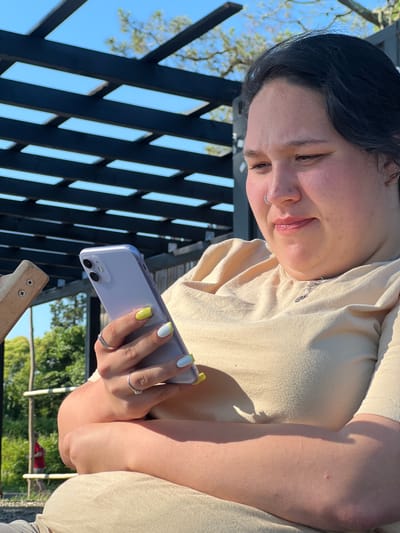 The image shows a young woman outdoors, holding a smartphone. She has dark hair and is wearing a light-colored shirt. She is looking down at her phone with a slight smile. Her nails are painted with a yellow and white design, and she is wearing two rings on her left hand. The background includes a wooden structure, greenery, and a bright blue sky, suggesting it is daytime and the weather is pleasant. The overall mood appears relaxed and casual. The setting is outdoors, possibly a patio or garden area.