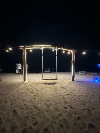 This image depicts a nighttime scene on a sandy beach in Jungutbatu, Indonesia. Centrally positioned on the light-colored sand is a rustic wooden swing structure, featuring two thick vertical posts and a horizontal beam. The entire structure is wrapped with bright white LED strip lights, and string lights with visible bulbs extend horizontally from the beam. An empty wooden swing with a dark seat hangs from two ropes. Text is partially visible on the horizontal beam, appearing to read "LURAKU".
To the left of the swing structure, a group of approximately three to four individuals stands on the sand; one person wears a white dress or skirt, and another a white t-shirt with dark shorts. In the background, nearer the water's edge, two to three figures are faintly visible, appearing to be seated or lounging. To the right of the swing, a small, light-colored table with a bottle is situated on the sand, and further right, a portion of a glowing blue neon-like art installation is discernible. The immediate foreground shows numerous footprints in the sand. The background consists of dark water under a dark night sky, with the illuminated swing structure serving as the primary light source for the foreground.