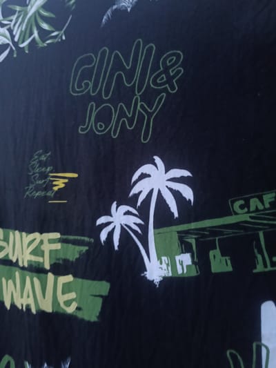 The image is a close-up of a black fabric with a vibrant graphic print. The graphic features text and illustrations in green, yellow, and white. At the top, the text "GINI & JONY" is written in a stylized green font. Below this, on the left side, is text that appears to read "Eat. Sleep. Surf. Repeat." with a yellow graphic element resembling waves. To the right of this, there are illustrations of palm trees in white. Further down, in larger font, the word "SURF" is printed in yellow, followed by "WAVE" in yellow below it, both overlaying green brushstroke-like shapes. On the right side of the fabric, there is a green and white illustration of what looks like a building or structure with the word "CAF" visible in green within a rectangular box. The image does not show any elements that would place it in a specific city like Altinkul, Uzbekistan, or any outdoor environment. It is solely a depiction of a printed textile.
