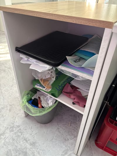 This is a top-down, eye-level shot inside a white shelf unit. The shelf is filled with various items, including folders, papers, plastic bags, and a red object in the upper right corner. A black crocodile-textured folder is on the left, with white papers sticking out. To its right are blue and light blue folders, and a plastic bag containing papers with the text "KOHTE" visible. Further to the right, a green package of paper is visible, labeled "STORA MultiCopy". A pink cloth is also present, along with a green plastic bag containing crumpled paper and a packet of "Coffee Deluxe". The shelf unit itself appears to be made of a light-colored wood on the left side and white material for the shelves. The background suggests a lightly textured, possibly marble-patterned floor. The lighting indicates an indoor setting, possibly during the daytime. No people are visible in the image.