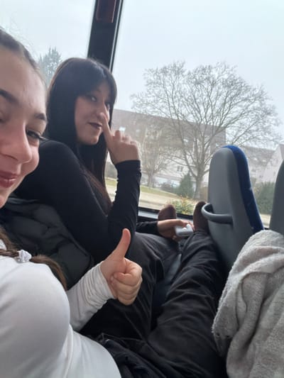 Three young women are in what appears to be a bus. One woman is in the foreground, smiling slightly, with dark lipstick. The woman behind her is wearing a black long-sleeved shirt and making a funny face. A child behind her is giving a thumbs-up. A building and tree are visible through the window. Someone's bare feet and a mobile phone are visible propped up against the seat in front.