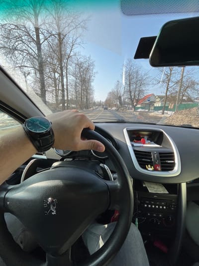 The scene is depicted from the interior of a Peugeot vehicle, specifically from the driver's perspective, looking forward through the windshield on a clear, sunny day in Maksimkovo, Russia. The driver's left hand, adorned with a large black-strapped wristwatch featuring a dark face and light green markers, is positioned on the black steering wheel. The dashboard presents an air vent system, a top-mounted console containing a prominent red object and other minor items, and below, the vehicle's integrated audio/climate controls with a paper slip inserted. A small, yellow, winged figurine is affixed to the right central air vent. The upper right portion of the windshield reveals a rearview mirror and a windshield-mounted device bracket. Outside the vehicle, a two-lane asphalt road extends into the distance under a clear, bright blue sky. Both sides of the road are lined with numerous tall, leafless deciduous trees. Accumulations of dirty snow are visible along the road shoulders. On the right side of the roadway, a white residential structure with a red roof is situated behind a blue-roofed shelter, consistent with a bus stop. Utility poles with overhead wires are present on the right. Distant vehicles are discernible on the road ahead.