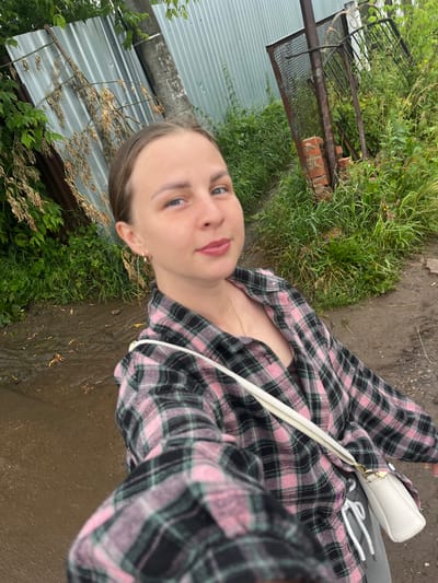 A young woman with light brown hair, pulled back, is taking a selfie outdoors. She is wearing a pink and black plaid flannel shirt over a dark-colored top and gray sweatpants. A small white crossbody bag is slung over her right shoulder. She is standing on a muddy path, with puddles of water visible. To her left, there is a corrugated metal fence and lush green foliage. To her right, there is overgrown grass, a rusty metal gate supported by brick, and a wooden pole. The scene suggests an outdoor location in a rural or semi-rural area, potentially the outskirts of Balezino, Russia, given the context. The weather appears overcast.