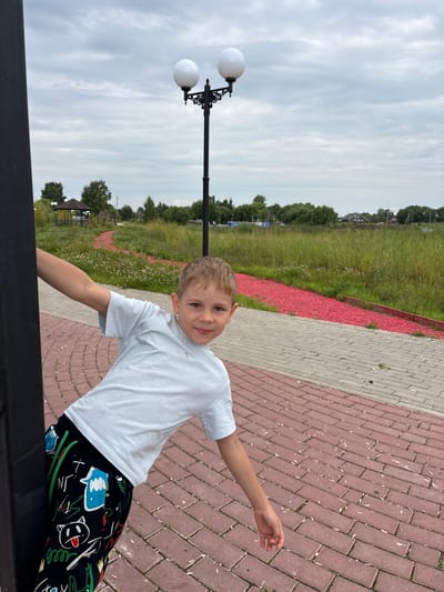 A young boy with blonde hair is posing for a photo in an outdoor setting. He is wearing a white t-shirt and black pants with colorful cartoon graphics. The boy is standing on a paved walkway made of red and gray bricks. To his left, there is a grassy area with scattered wildflowers and a lamppost with two white globes. In the background, there are trees, a small gazebo, and some houses. The sky is overcast. The overall mood of the image is casual and cheerful. The photo appears to have been taken during the daytime. The location is identified as Breytovo, Russia.
