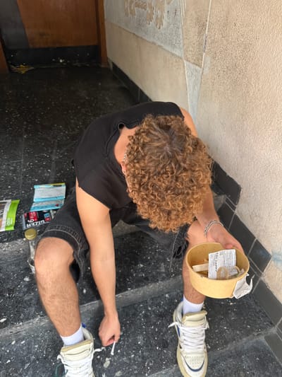 The image shows a young person with curly hair sitting on steps outdoors. They are wearing a black t-shirt and denim shorts, with white sneakers and socks. The person is holding a small paper bowl in their right hand, which contains what appears to be receipts and a small packet. Their left hand is near their left foot, holding a small white object, possibly a cigarette or similar item. On the steps to the left of the person, there are several items, including what looks like rolling papers, a bottle, and a green packet. The environment appears to be an urban setting with a speckled dark floor and a textured wall. No specific landmarks in the city of Hamm, Germany are visible.