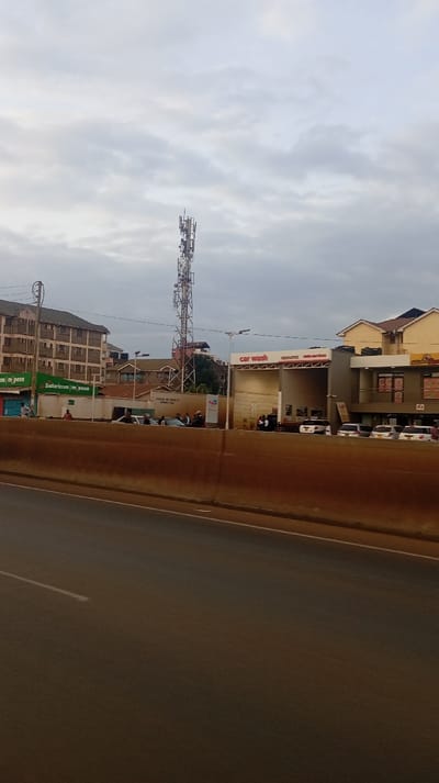 A street view captures a car wash with parked cars and individuals standing outside. A tall telecommunications tower rises behind the building. Adjacent structures include a building with "Safaricom M-pesa" signage. The sky is overcast, creating a diffused light. A road with a median barrier runs in the foreground.