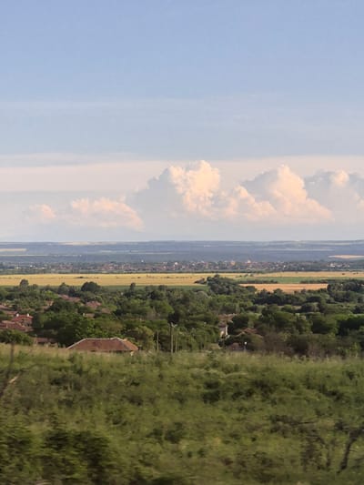This is a medium-distance, vertical, eye-level shot of a landscape that appears to be in or around Kozar Belene, Bulgaria. The image captures a hazy, overcast sky with large, puffy white clouds. Below the sky, a distant, flat landscape unfolds, featuring agricultural fields of yellow and green, interspersed with scattered buildings and a faint hint of a town or city in the middle ground. In the foreground, a densely forested area with numerous red-roofed houses is visible. The overall scene is one of rural settlement surrounded by cultivated land and natural greenery. There are no people or animals depicted in the image.