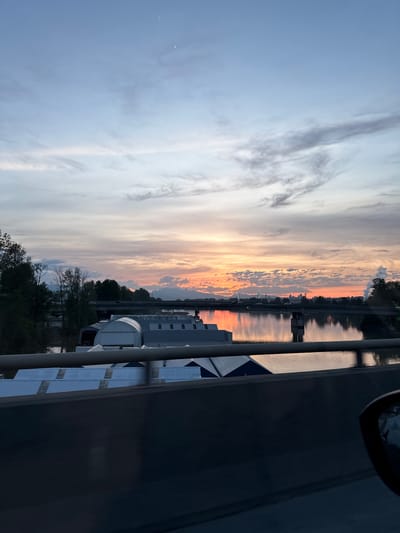 The image displays a tranquil sunset scene over a body of water, likely a river or strait, in Delta, Canada. The sky dominates the upper portion, transitioning from a soft light blue and gray on the left, with wispy white and gray clouds, to vibrant hues of orange, pink, and yellow near the horizon on the right, indicating the setting sun.

Below the sky, the calm water perfectly mirrors these vivid sunset colors, creating a striking reflection across the middle of the frame. Along the distant horizon, a silhouette of a treed shoreline and a few indistinct structures can be observed.

In the midground on the left, a line of trees with varying foliage stands by the water's edge. To the right, several modern buildings with white or light-colored exteriors are situated along the waterfront. These structures feature distinctive architectural elements, including arched roofs and what appear to be solar panels.

The immediate foreground on the right is largely occupied by a dark, angular structure, possibly part of a bridge, a large vehicle, or an adjacent building, which partially obscures the view of the water and buildings. A dark metal railing runs horizontally across this foreground element. A small, partial circular object is visible at the very top right corner.

No people are visible, and there are no specific activities taking place, contributing to the peaceful atmosphere. The scene is illuminated by the soft, warm light characteristic of dusk. There is no visible text in the image.