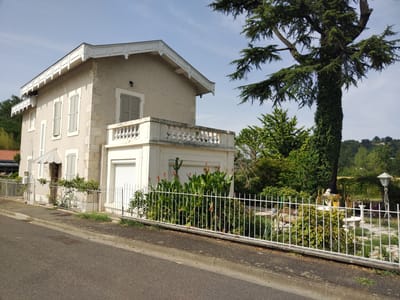 The image depicts a two-story house with a light-colored stucco exterior, located on a street in Le Passage, France. The house has white trim around the windows and a decorative white balcony railing on the upper floor. A light-colored metal fence runs along the front of the property, separating it from the paved street. Lush green vegetation, including flowering plants and trees, surrounds the house and fills the yard. A tall, mature tree with a thick trunk covered in ivy stands prominently to the right of the house. The sky is clear and blue with a few wispy clouds. The scene appears to be a residential area on a sunny day.