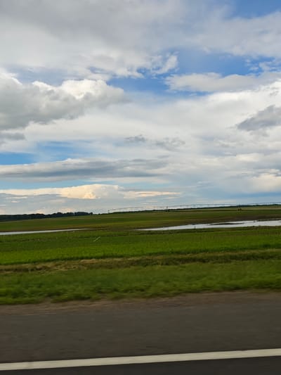 This media file captures a wide shot of a rural agricultural landscape under a dynamic sky. The scene is dominated by vast, lush green fields, some appearing to be flooded or irrigated, with distinct rows indicating cultivation. In the middle ground, a long, metallic irrigation system with numerous sprinklers stretches across the horizon, suggesting active farming operations. A line of dark green trees forms a distant boundary to the fields. The sky is filled with a dramatic interplay of white and gray clouds, with patches of bright blue visible, indicating a day with varied weather. The lighting suggests it could be late afternoon or early evening, with diffused sunlight. The foreground features a dark asphalt road with a prominent white line, suggesting the vantage point is from a moving vehicle. The overall impression is one of agricultural activity and the expansive, open nature of the countryside. No people are clearly visible. The location context is Красненскі сельскі Савет, Belarus.