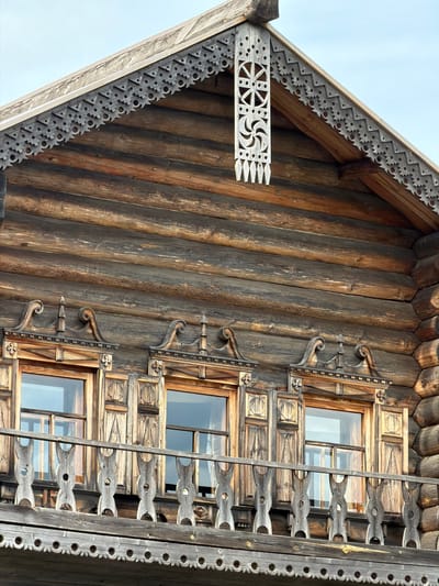 The image displays a close-up view of the upper section of a traditional wooden log house in Dnp Ryzhiki, Russia. The house features intricate, carved wooden trim along the eaves of the roof and around the windows. The walls are constructed from horizontally stacked, rough-hewn logs. There are three windows visible, each with wooden shutters and ornate frames. Below the windows, a wooden balcony railing with carved detailing runs across the facade. The sky is visible in the upper portion of the image. No people are present. The overall impression is one of traditional Russian architecture and craftsmanship.