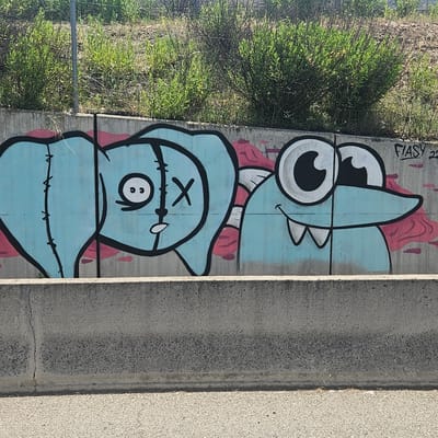 The image depicts a section of a concrete barrier, likely located near a highway in the city of Alcalá de Henares, Spain. The barrier is painted with graffiti featuring two cartoonish blue figures outlined in black. The figure on the left has a stitched-together appearance, with one eye depicted as two small dots and the other as an "X." The figure on the right has large, wide eyes and two prominent teeth. Red accents are painted around the figures. A signature "FIASY 22" is visible in the upper right corner of the concrete wall. Vegetation grows above the barrier. The ground in front of the barrier is paved.