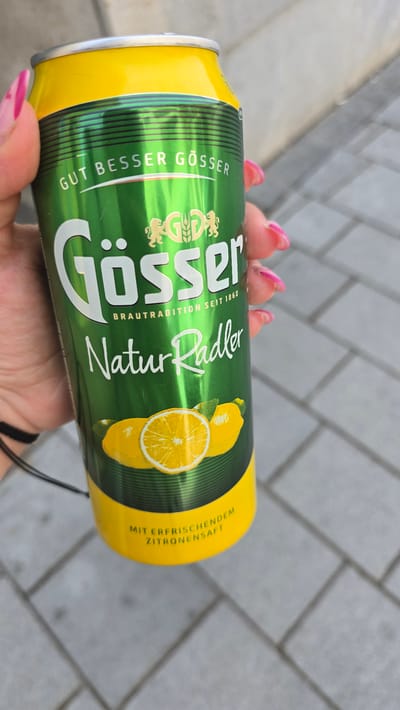 A person is holding a can of Gösser Natur Radler lemon beer. The can is green and yellow, with lemons printed on the side. The person has pink nails. The background is a grey brick surface. The location is not discernible from the image and cannot be determined to be in Zirndorf, Germany.