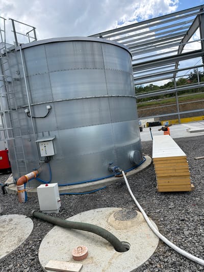 The image depicts an outdoor construction site in Jodłownik, Poland, dominated by a large, cylindrical metal tank. The tank is grey, with visible rivets and seams, and has a ladder leading up its side. Adjacent to the tank, a steel structure for a building is under construction, with exposed beams and supports. Several construction materials are present, including a stack of yellow and white insulated panels and a red generator. Various pipes and hoses are connected to the tank and extend to the ground, where they connect to concrete manhole covers and other underground infrastructure. A worker in an orange vest and blue hard hat is visible in the middle ground, carrying a piece of wood. The ground is covered with grey gravel. The sky is partly cloudy.
