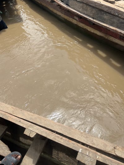 An aerial shot shows a section of the River Benue in Nemnei, Nigeria, with two wooden boats docked alongside each other. The water is a muddy brown, with ripples and sunlight reflecting off its surface. The boats appear weathered, with visible planks and aged wood. In the foreground, a person's foot wearing a black sandal is resting on the edge of one of the boats. A person wearing dark clothing is partially visible in the upper left corner of the image, seemingly standing in or near the water. The scene suggests a typical day on the river, with boats and human activity.