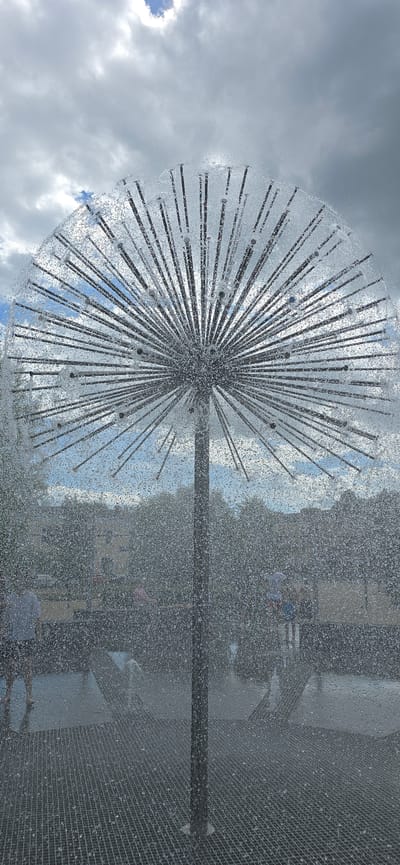 The image shows a dandelion-shaped water fountain in motion, with numerous fine water jets creating a sphere of mist and droplets. The fountain is supported by a central dark pole. The background reveals a cloudy sky with patches of blue, and behind the fountain, there are blurred trees and buildings, suggesting an outdoor urban setting. In the lower half of the image, several figures of people are visible, some of whom appear to be children, interacting with the water feature. The ground beneath the fountain is a metal grate, likely designed for water drainage. The scene captures a dynamic and playful moment in what appears to be a public space or park, possibly in the city of Bryansk, Russia, given the prompt. The overall impression is one of summer recreation and a refreshing atmosphere created by the water fountain.