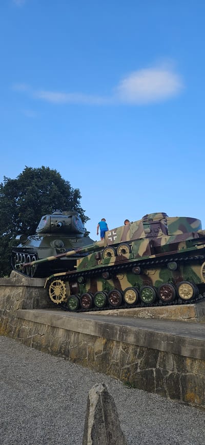 The image captures an outdoor scene with two military tanks displayed on a stone embankment under a clear blue sky. In the foreground, a camouflaged tank, featuring shades of green, brown, and black, is prominently positioned. A person in a blue shirt is standing on top of this tank, facing away from the camera. To the left, a dark green tank with a red star emblem is partially visible, positioned higher on the embankment. A large tree with dense foliage is situated behind the dark green tank. The ground in front of the embankment is covered with gravel. The scene is located at the Dukla Pass Memorial in Svidník, Slovakia, a significant historical site commemorating the Carpathian-Dukla Operation during World War II.