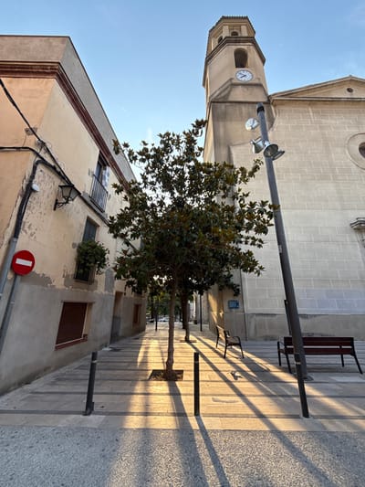 The image shows a street scene in La Pobla De Claramunt, Spain. On the left is a multi-story building with a cream-colored facade, some of which is peeling. A red and white "no entry" sign is visible on a pole in front of the building. A street lamp is attached to the building's exterior. There are windows on the building, some with shutters and one with a hanging plant. The building is adjacent to a sidewalk paved with light-colored square tiles and darker rectangular accents. A young tree is planted in the middle of the sidewalk, casting long shadows. Beyond the tree, there are two park benches. To the right of the sidewalk is a large, light-colored stone building with a clock tower. The clock face shows approximately 11:45. The sky is clear and blue. The sun is low in the sky, casting long shadows across the scene, indicating it is likely early morning or late afternoon.