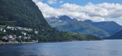 The image depicts a scenic view of a residential area nestled on a steep, tree-covered mountainside overlooking a body of water. The architecture consists primarily of white and light-colored houses with red roofs, typical of Norwegian villages. The settlement appears to be situated in the city of Volda, Norway, a location known for its dramatic fjords and mountainous terrain. The water in the foreground is calm, reflecting the cloudy sky and the surrounding landscape. In the background, imposing mountains rise, some with snow-capped peaks, indicating a high latitude or elevation. The overall impression is one of tranquility and natural beauty, characteristic of the Norwegian countryside. There are no visible people or animals in the scene, and the environment appears undisturbed.