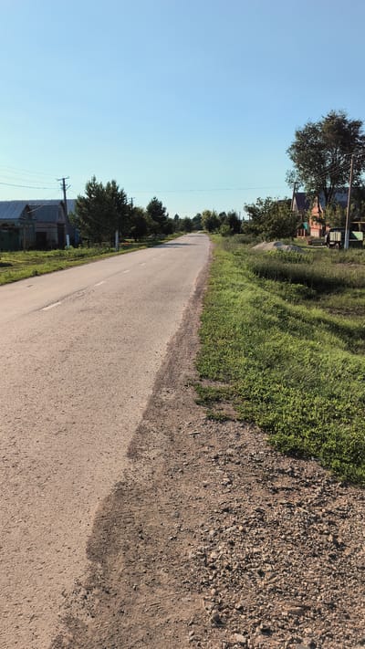 The image depicts a rural road in Alkyino, Russia, under a clear blue sky, suggesting fair weather and daytime. The road, paved and slightly worn, curves gently to the right, bordered by grassy verges and patches of gravel and dirt. On the left side of the road, a cluster of houses with traditional architecture, some featuring blue trim and corrugated roofs, are visible amidst trees. Power lines and poles run alongside the road, hinting at the presence of utilities in the area.
On the right side of the road, more greenery dominates, with lush grass and trees. Further down the road on this side, a few more houses are visible, one appearing to be brick with a reddish hue. A truck with a green trailer is parked near one of the houses. The overall scene conveys a sense of quiet rural life. The lighting suggests it is a sunny day, likely morning or afternoon. There is no visible text in the image.