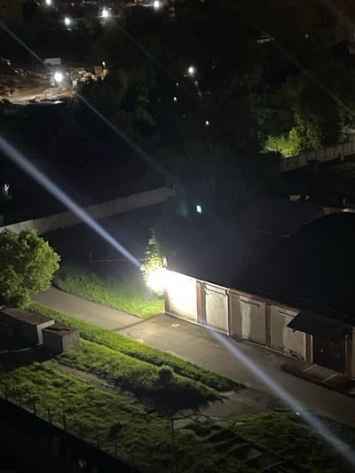 An exterior nighttime aerial shot, captured in Tarasovka, Russia, showcases an urban industrial or storage area. The scene is illuminated by multiple artificial light sources, casting long beams and bright spotlights across the landscape. Dominating the foreground is a long, low building, likely a garage or storage facility, with a dark, sloped roof and a row of light-colored doors, each framed by red trim. One section of this building is intensely lit from within, suggesting an active light source or an open area. Adjacent to the building, a paved road or pathway runs diagonally, bordered by a strip of green, unkempt grass. Further to the left, a large, lush green tree stands prominently, with a rough, uneven grassy area and two concrete structures, possibly storage bins or small buildings, situated behind it. In the background, the landscape becomes darker and more obscured, with more indistinct structures and patches of light visible amidst dense trees and foliage, suggesting a more remote or undeveloped area. The overall impression is one of an active, albeit dimly lit, industrial or storage zone at night.