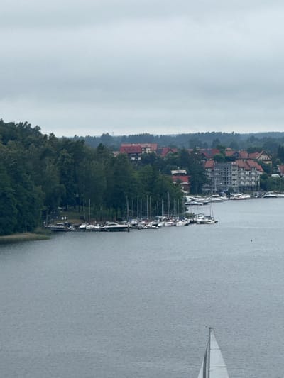 The image depicts a serene lakefront scene in Mikołajki, Poland. In the foreground, a large body of water occupies the majority of the frame, with a small, white sailboat with its sail raised visible in the lower right corner, heading away from the viewer. The middle ground showcases a bustling marina filled with numerous moored yachts and sailboats. Beyond the marina, a dense line of green trees forms a natural backdrop. Further inland, several buildings with red-tiled roofs and at least one multi-story, grey structure are nestled among the trees, suggesting a developed lakeside community. The sky is overcast with a light grey hue, casting a diffused light over the entire scene.