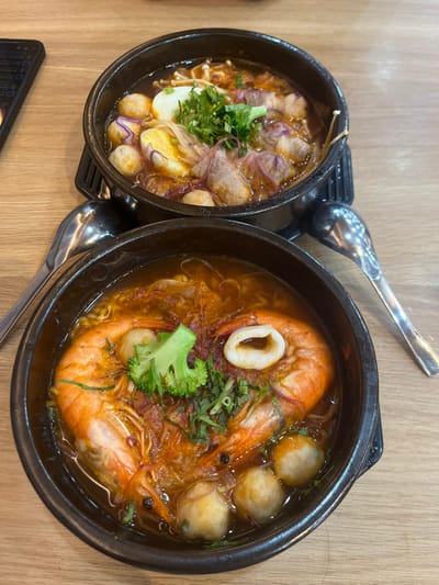 The image displays a close-up, elevated view of two individual servings of hot Vietnamese noodle soup dishes, presented on a light-colored wooden table in the city of Cần Thơ, Vietnam.
The upper dish, housed in a black ceramic bowl on a black base, contains a reddish-brown broth. Ingredients visible include sliced meat, several white meatballs, halved hard-boiled eggs, enoki mushrooms, shredded purple cabbage, and chopped green herbs as garnish.
The lower dish, also in a black ceramic bowl on a black base, features a reddish-orange broth. This dish contains two large, cooked prawns, several white meatballs, one prominent white calamari ring, a small green broccoli floret, and chopped green herbs. Fine red strands are distributed throughout the broth.
Two silver-colored metal spoons are present: one to the left, positioned between the two bowls, and another to the right, adjacent to the lower bowl. A dark-colored smartphone is partially visible in the upper-left corner of the frame, resting on the wooden table surface.